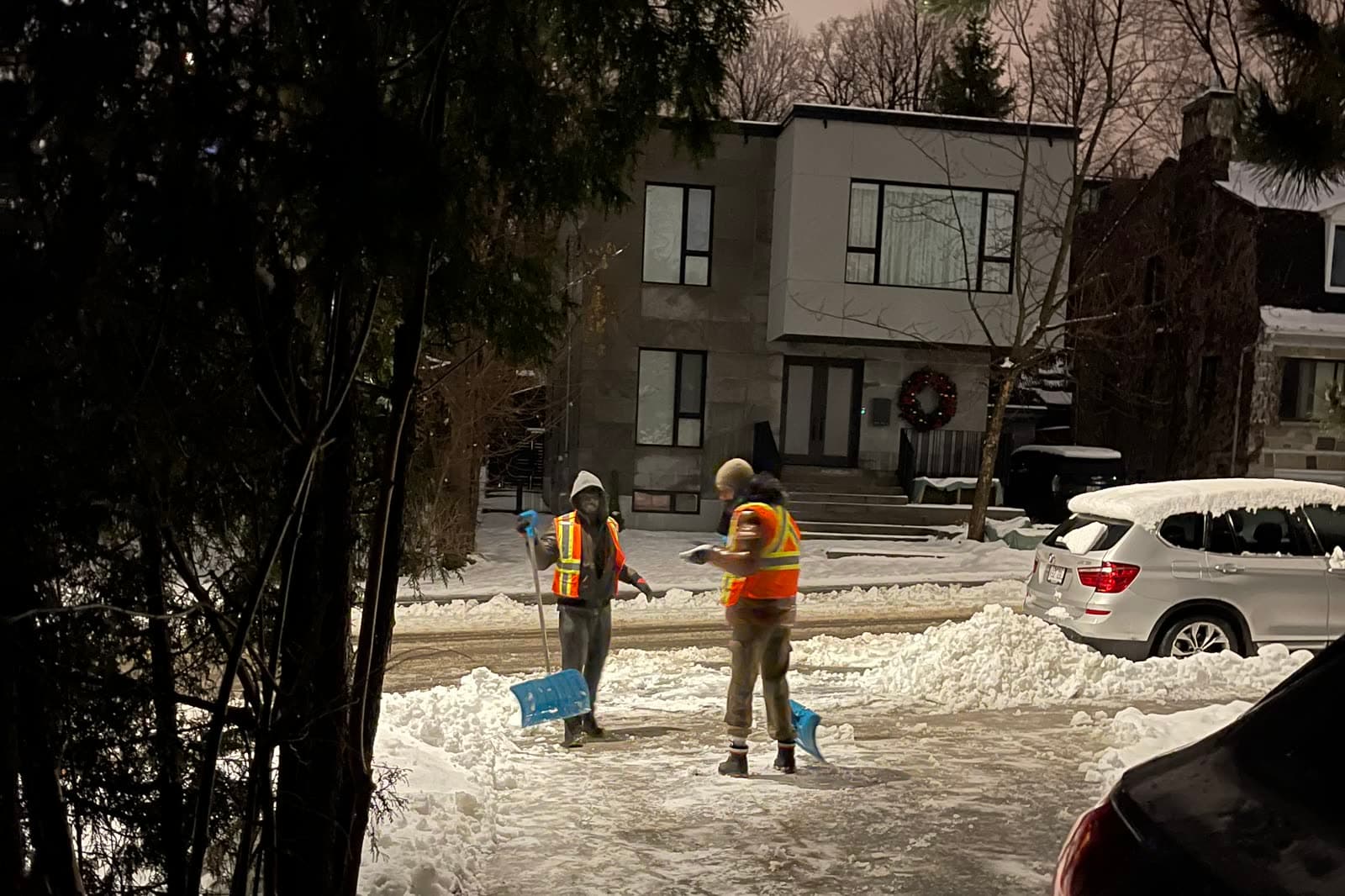 Two Snoen crew members in high-visibility vests clearing a residential driveway at night after a snowfall